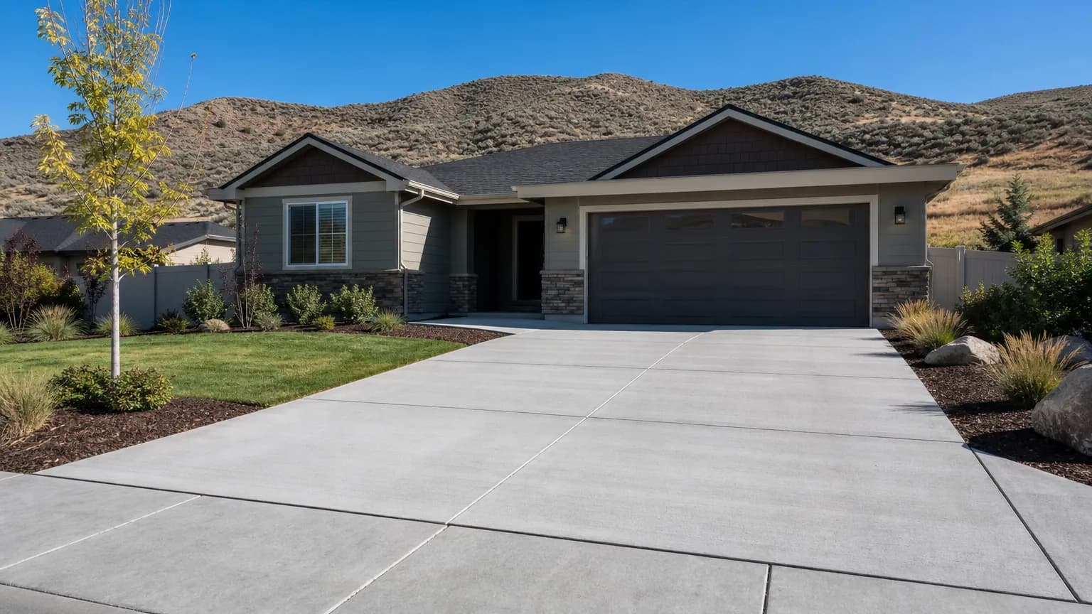 New light gray broom-finish concrete driveway with saw-cut joints in front of a single-story home, with sagebrush foothills behind