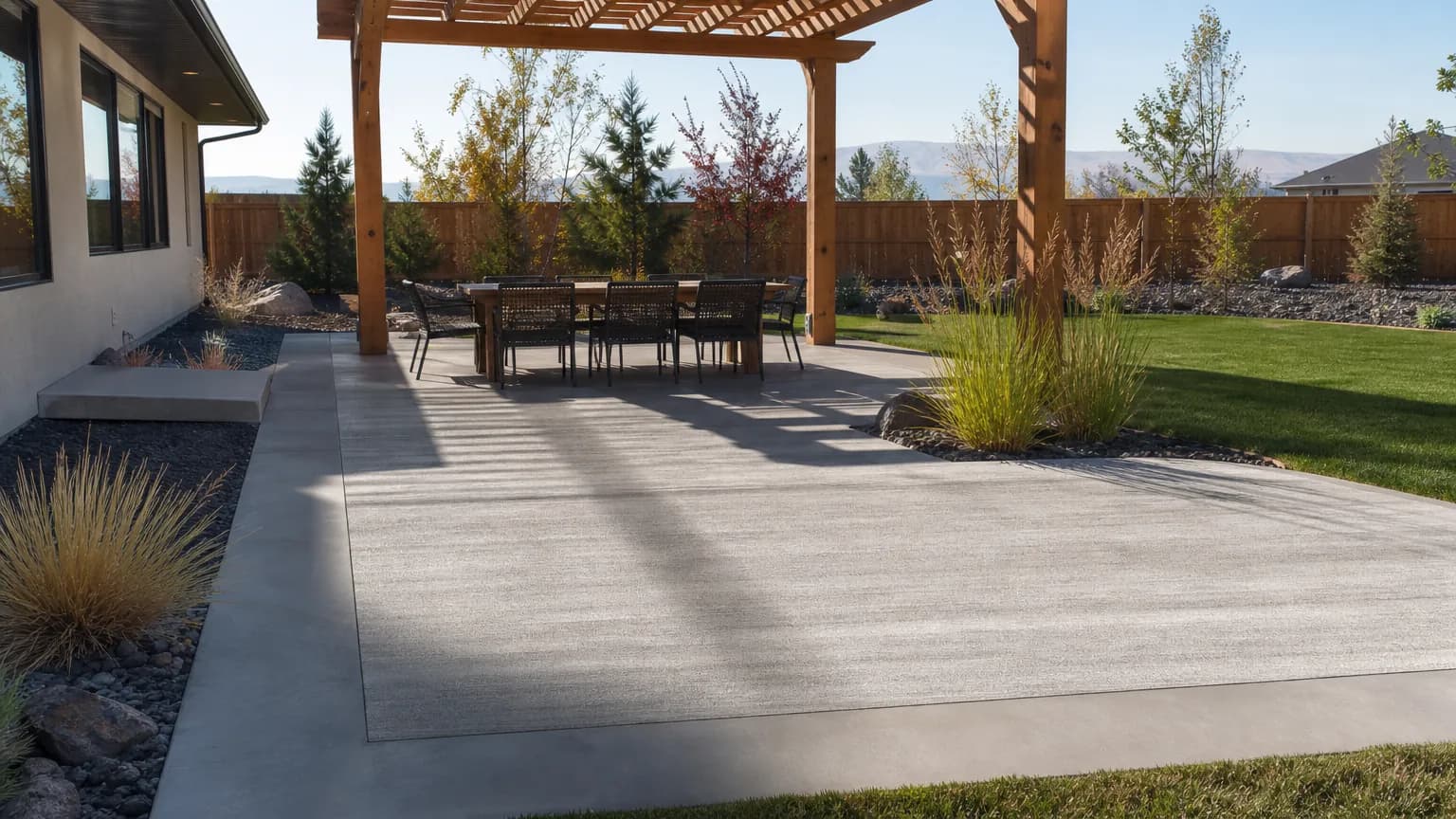 Backyard concrete patio with a troweled border and broom-finish center under a wood pergola, with ornamental grasses and lava rock